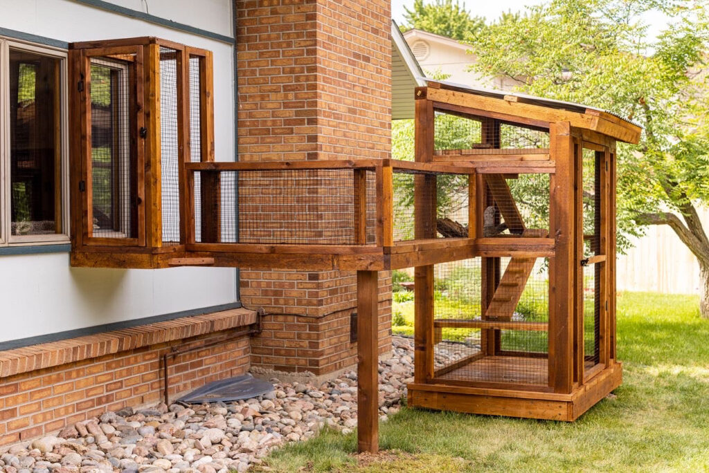 A wooden catio with a connecting tunnel attached to a house, featuring wire mesh and multiple levels, situated in a backyard with grass, trees, and brick elements.