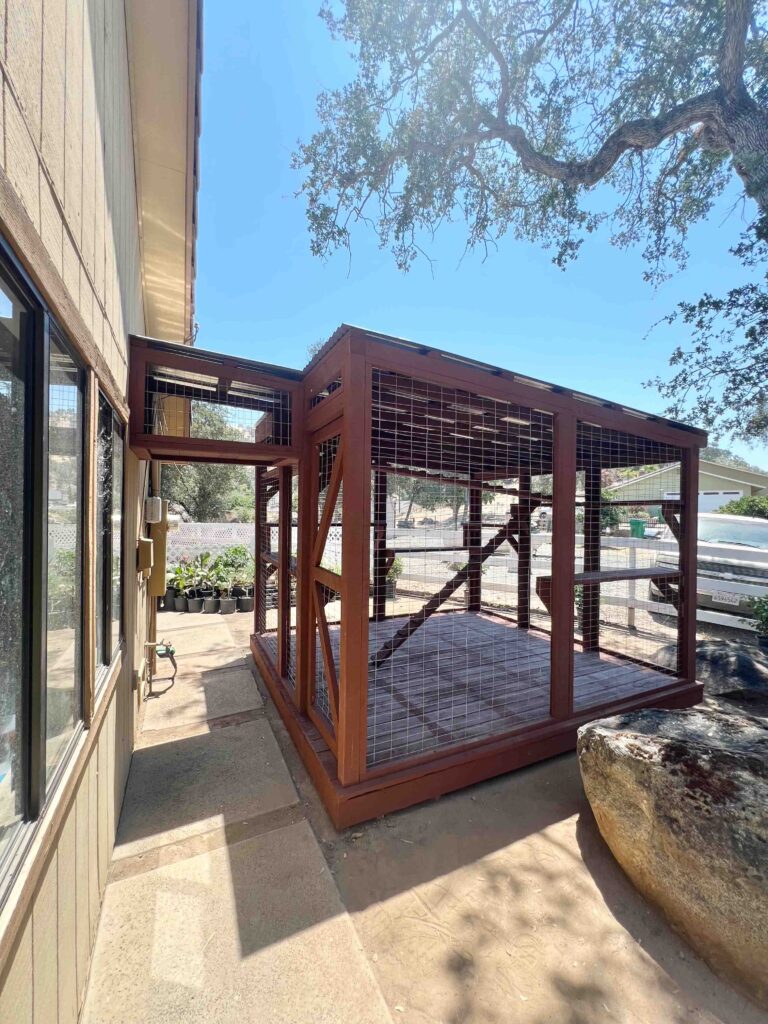 A custom-built wooden catio attached to the side of a house, featuring a sturdy frame, wire mesh walls, and a connected tunnel leading to the house. The catio sits on a concrete pathway with outdoor landscaping visible in the background.