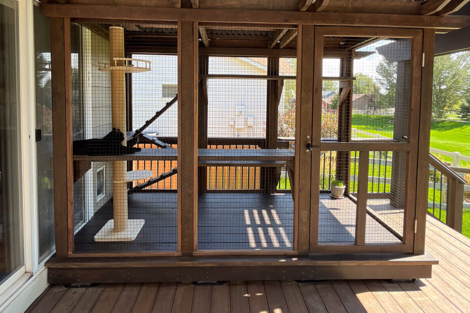 A custom-built wooden catio with mesh walls, attached to a house. Inside, a black cat lounges on a platform next to a tall scratching post, while multiple shelves and ramps provide climbing opportunities.