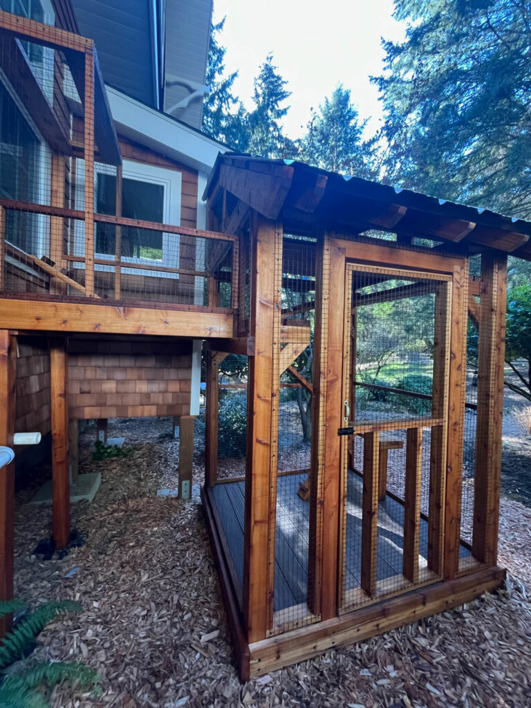 A well-constructed wooden catio attached to a house, featuring a secure wire mesh enclosure, a covered roof, and an elevated walkway leading from a window to the main outdoor area.