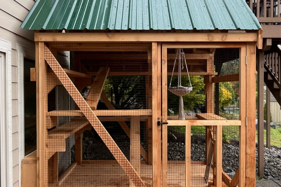 A compact yet spacious wooden catio with a green metal roof is securely attached to the side of a house. The enclosure features multiple wooden shelves, ramps, and a cozy macramé hanging hammock for feline relaxation. Sturdy wire mesh ensures safety while allowing fresh air and outdoor views. A small cat door connects the catio to the house, providing easy indoor-outdoor access.